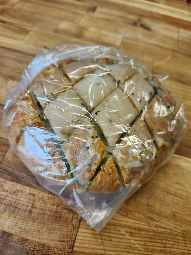 20260605_123106 1 | JustDane | Changing Lives. Changing the Community Round loaf of stuffed bread cut into square wedges and wrapped in plastic on a wooden surface.