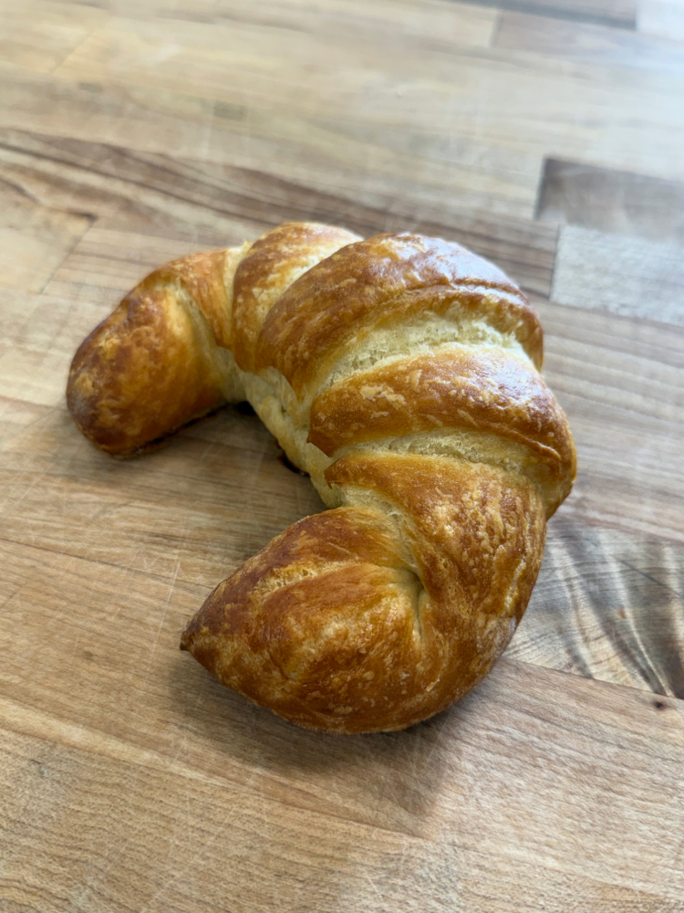 IMG_20260715_101111 1 | JustDane | Changing Lives. Changing the Community Golden-brown croissant on a wooden cutting board.