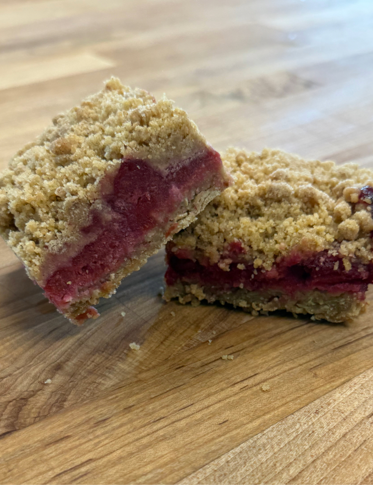 cherry crumble bar | JustDane | Changing Lives. Changing the Community Two berry crumble bars on a wooden cutting board, showing a red jam-filled center and a crumbly streusel topping.